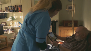 A nurse leans over to assist a man sitting in a living room, looking at a tablet.