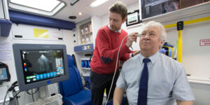 A man in a red sweater performs an ultrasound on an older man inside an ambulance equipped with medical devices.