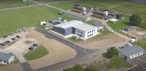 Aerial view of a white building with a parking lot and surrounding greenery and other structures.