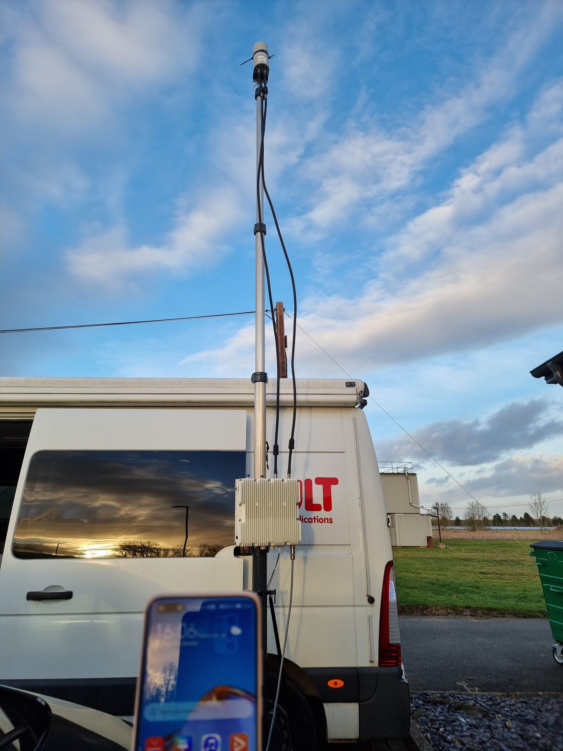 A white van with a mounted antenna, outdoor equipment, and a smartphone in the foreground at sunset.