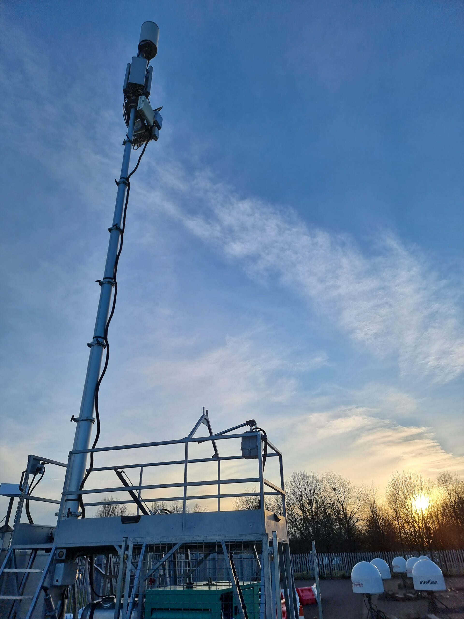 Tall metal pole with cameras on a platform, set against a blue sky at sunset, with trees and fencing below.