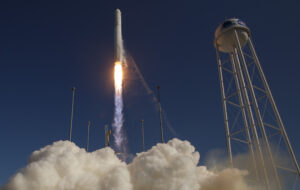 A rocket launches into a clear sky, with clouds of smoke below and a water tower nearby.