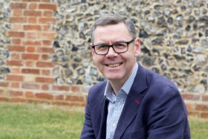 Smiling man wearing glasses and a blazer seated outdoors, with a brick and stone wall in the background.