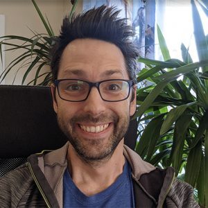 A smiling man with glasses and spiky hair sits in front of large green plants.