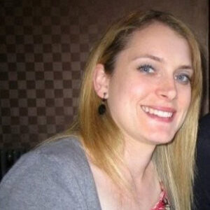 Smiling woman with long blonde hair wears a gray top, posing against a checkered background.