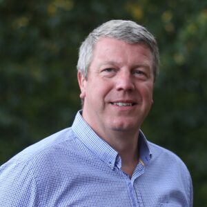 Smiling man in a blue checkered shirt stands outside with a blurred green background.