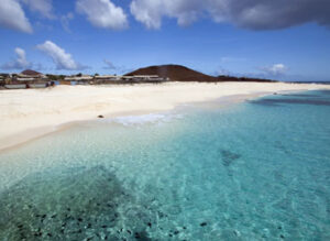 Clear turquoise waters and a sandy beach under a blue sky with scattered clouds. Buildings and a hill in the distance.