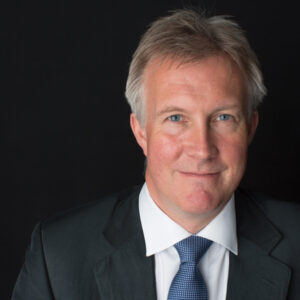 Smiling man in a suit with a blue tie against a dark background.