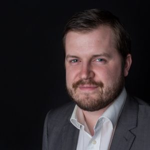 A man in a suit with a beard and short hair smiles against a dark background.
