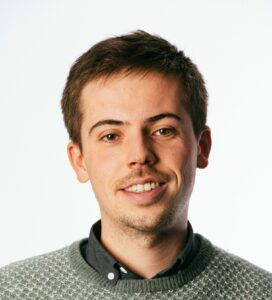 A man with short brown hair, wearing a gray sweater over a dark shirt, smiles against a plain white background.