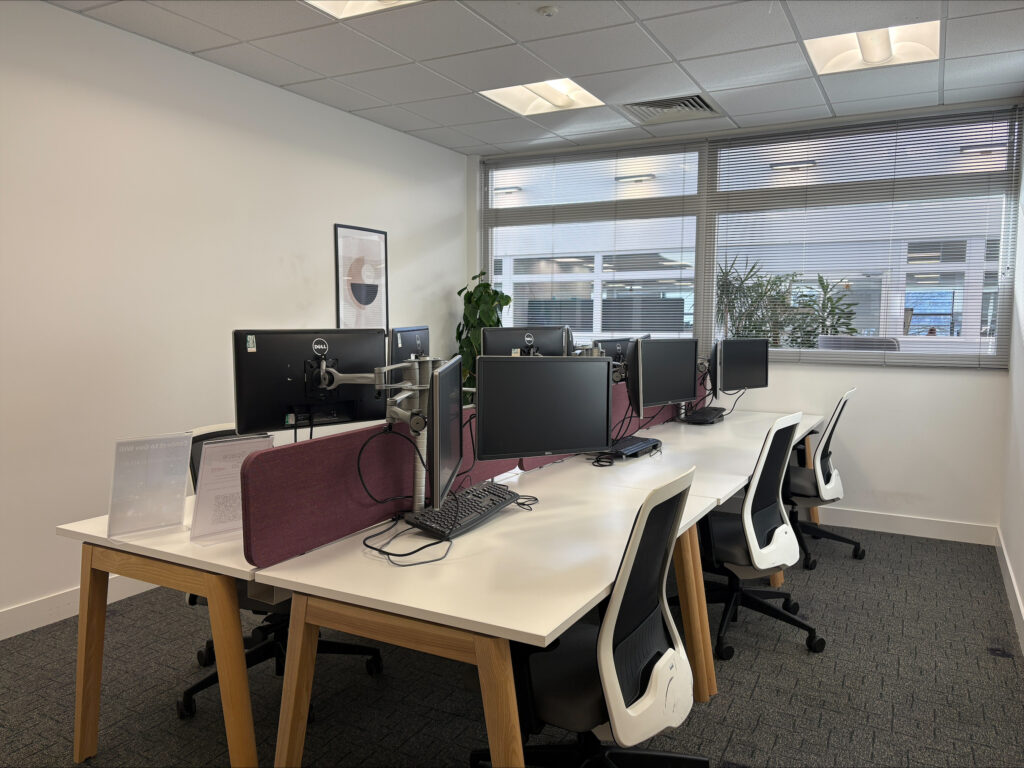 A modern office with four desks, computers, and chairs, next to large windows with blinds.