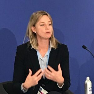 A person speaking on stage in business attire, gesturing with hands, against a blue background.
