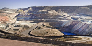 Open-pit mine with terraced rock layers, dirt roads, and a blue water pond on a clear day.