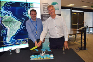 Two men cut a cake beside a screen showing a map, with banners in the background.