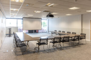 Empty conference room with U-shaped tables, chairs, a screen, projector, and a podium near large windows.