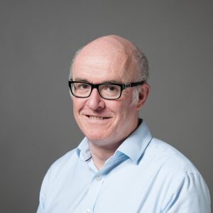 Smiling man in glasses and a light blue shirt against a gray background.