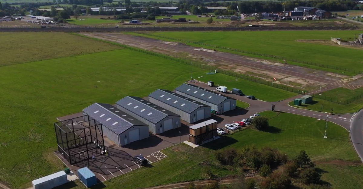 Aerial view of several industrial buildings by a rural road, surrounded by green fields and parked cars.