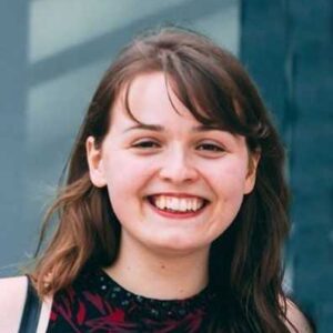 Young woman with long brown hair smiling warmly against an outdoor backdrop.
