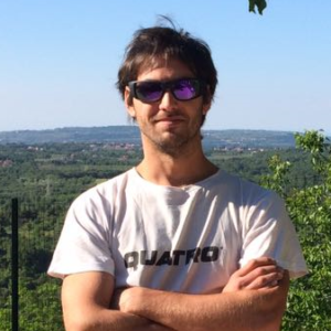 Man with sunglasses and a white shirt stands with arms crossed in front of a scenic green landscape.