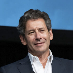 Man with gray hair in a dark suit and white shirt, smiling against a blue background.