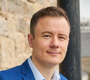 Person with short hair in a blue suit, standing outdoors against a stone wall, looking at the camera.
