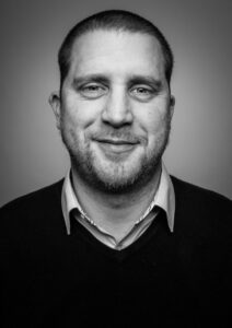 Man with short hair and a beard, wearing a collared shirt and dark sweater, smiling at the camera in grayscale.