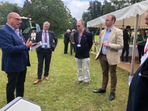 A group of people in business attire having a conversation outdoors near a white event tent on a grassy lawn.