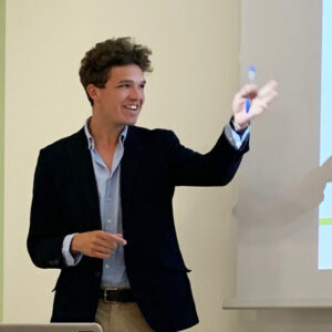 Person in a suit speaking, gesturing with one hand, holding a pen, standing near a projection screen.