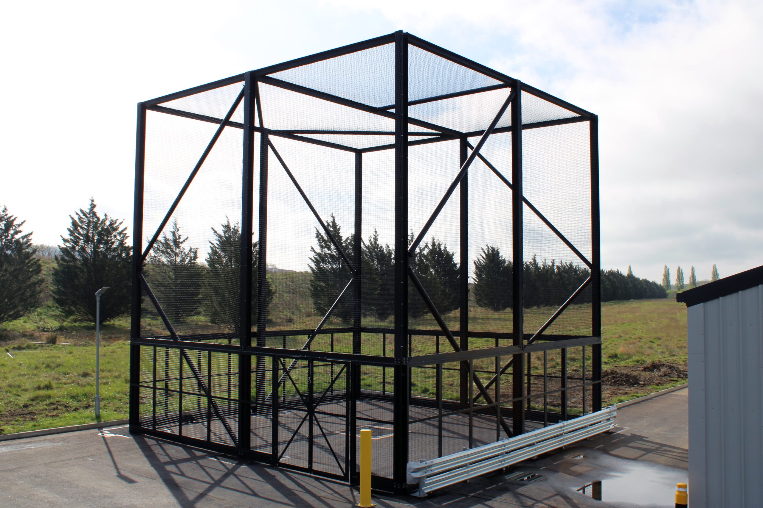 A large black metal cage structure stands outdoors on a paved area near grass and trees.