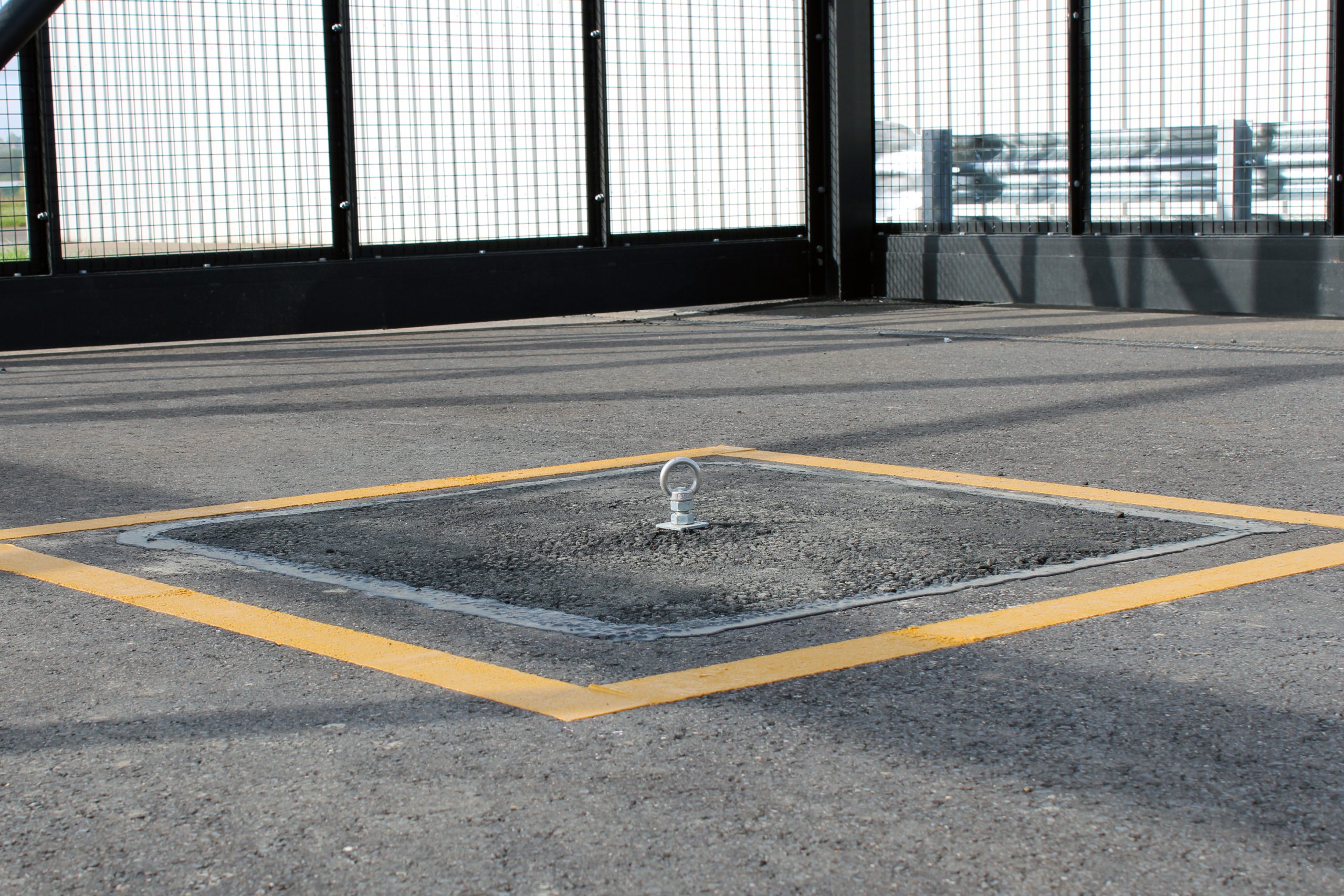 A small metal ring set in a square patch of asphalt outlined with yellow lines on a fenced pavement.