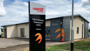 Sign and building of Future Networks Development Centre with logo, surrounded by cloudy sky and grass.
