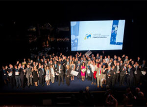 Large group of people on stage at an awards ceremony with a presentation screen in the background.