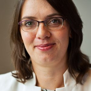 Person with glasses and dark brown hair, wearing a white shirt, smiles softly at the camera.