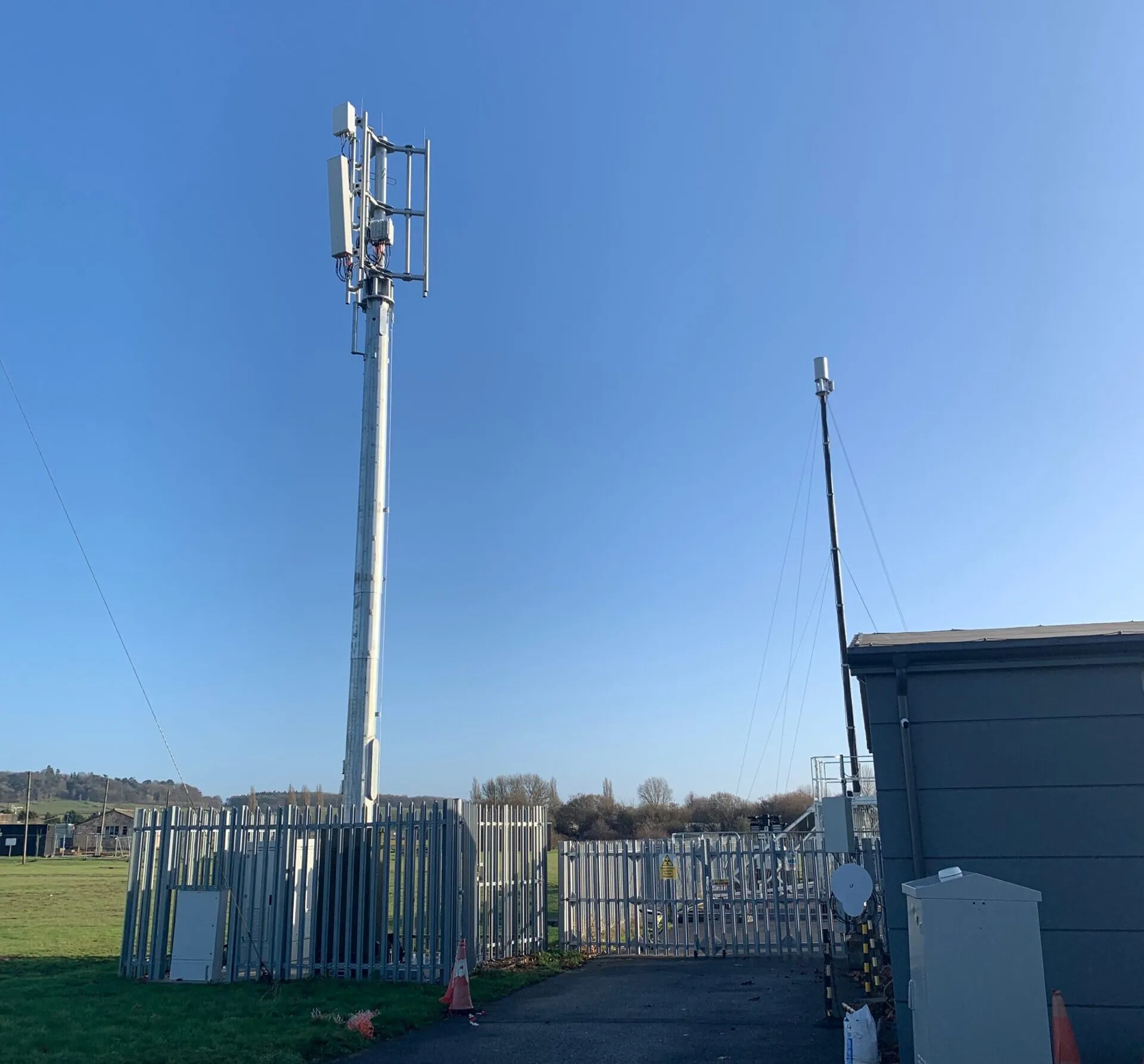 A cell tower and utility equipment stand inside a fenced area under a clear blue sky.