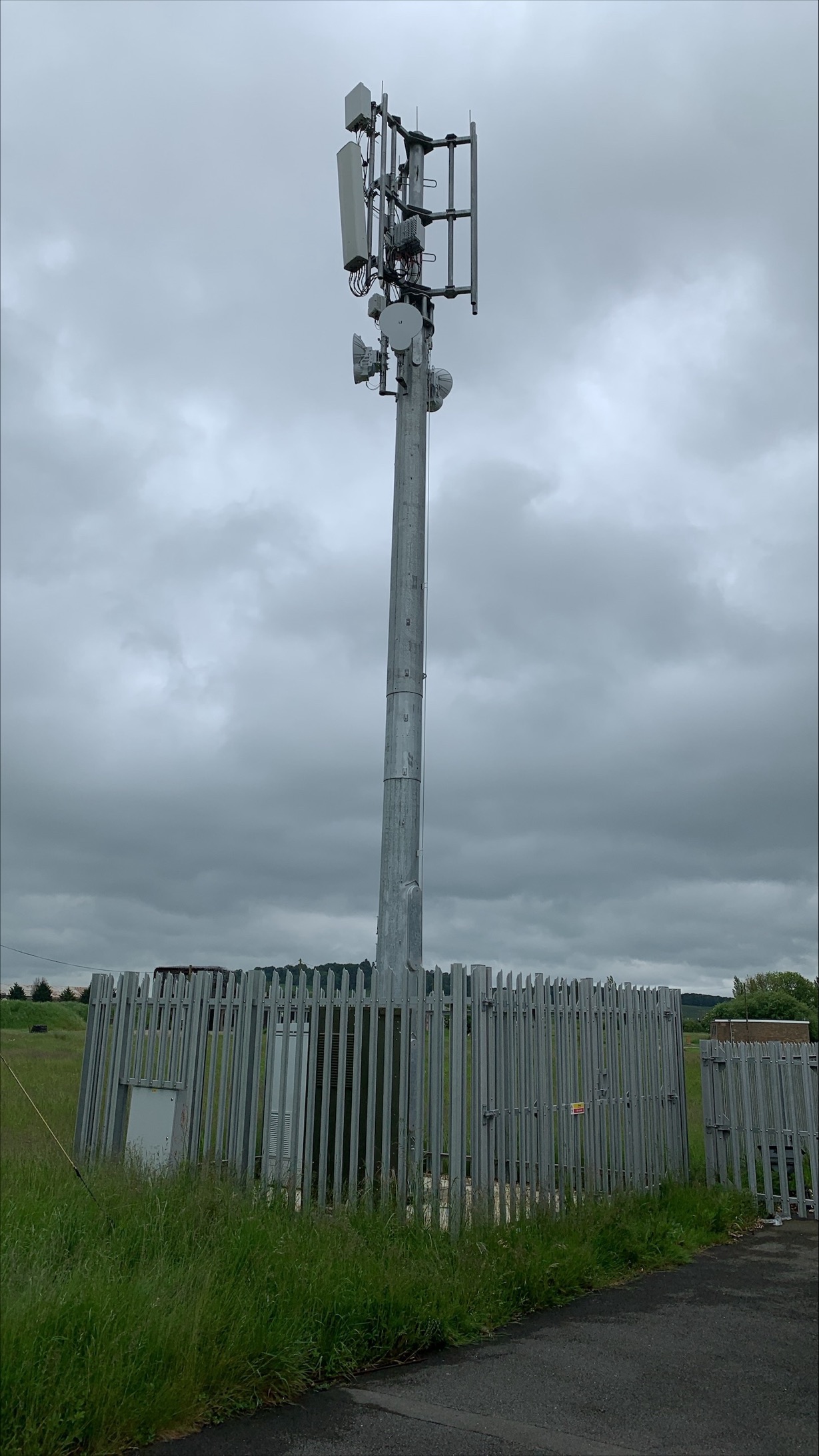 A tall cell tower stands inside a fenced area under a cloudy, overcast sky.
