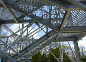 Metal staircase structure with crisscrossed beams and mesh, set against a backdrop of trees and a cloudy sky.