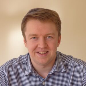 A person smiling and wearing a checkered shirt against a plain background.