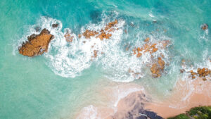 Aerial view of turquoise ocean waves crashing on rocky shoreline with sandy beach and greenery.