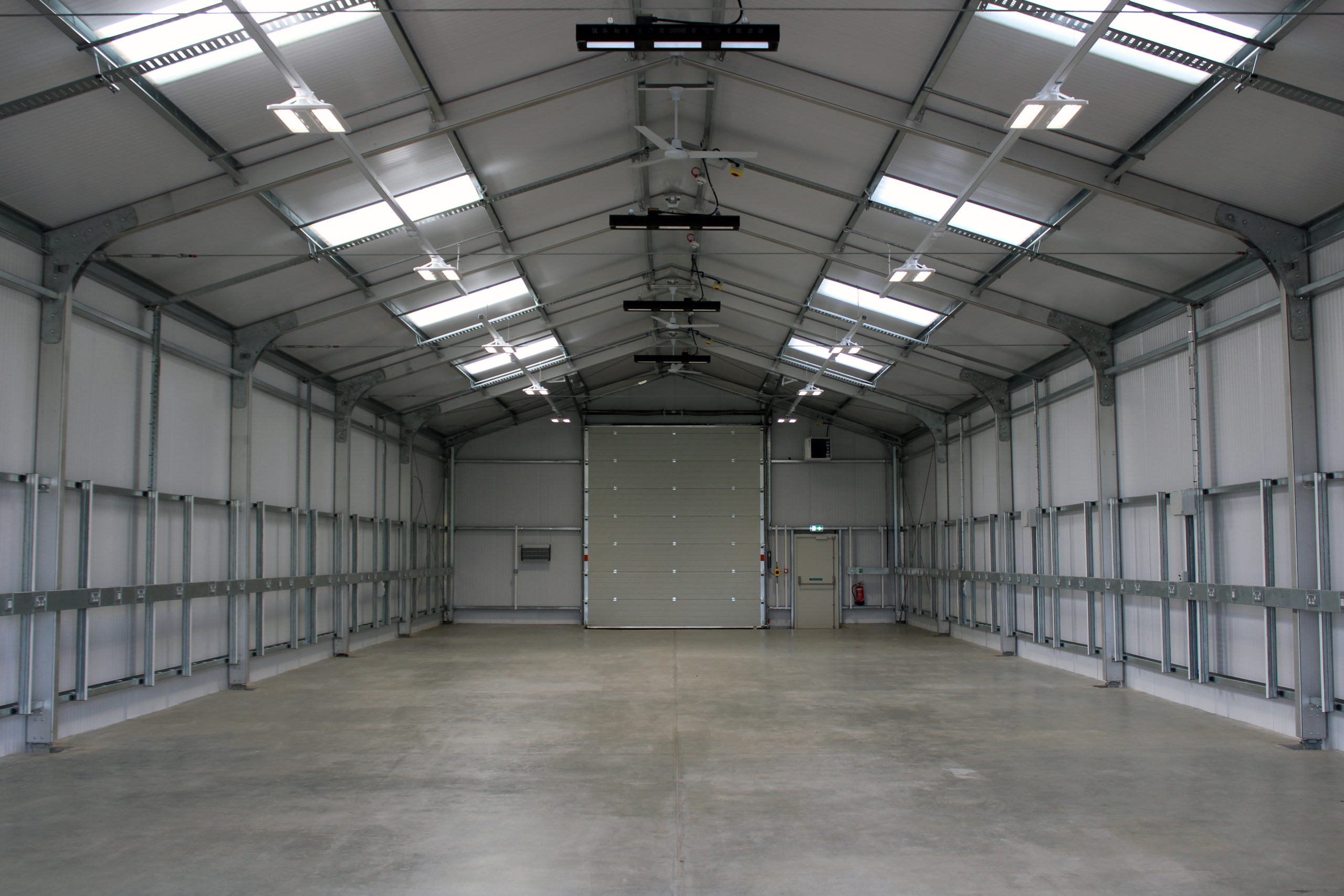 Spacious, empty warehouse with a high metal ceiling, skylights, and a large closed garage door at the end.
