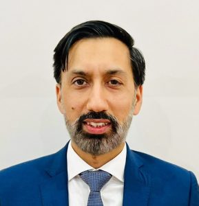 A man with dark hair and a beard wearing a blue suit and tie, smiling against a plain background.