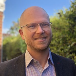 Smiling man with glasses, bald head, beard, and a suit, standing outdoors with greenery and a blue sky background.