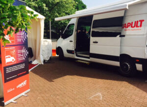 A white van with an open side door is parked on a brick road next to a tent and a sign with colorful graphics.