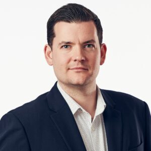 Man in a dark suit and white shirt poses against a light background.