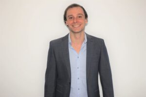 Smiling man in a gray suit and light blue shirt standing against a white background.
