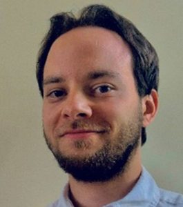 A man with short hair and a beard wearing a light-colored collared shirt smiles slightly against a plain background.