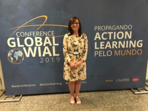 A person stands smiling in front of a "Global WIAL 2019" conference banner indoors.