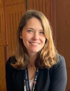Person with shoulder-length hair smiling, wearing a dark top and a lanyard, sitting indoors near wooden paneling.