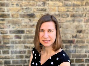 Person with shoulder-length brown hair, wearing a black polka dot dress, smiling in front of a brick wall.