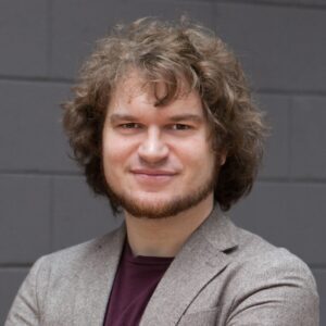 Person with curly hair and a beard, wearing a gray blazer, smiling in front of a dark gray wall.