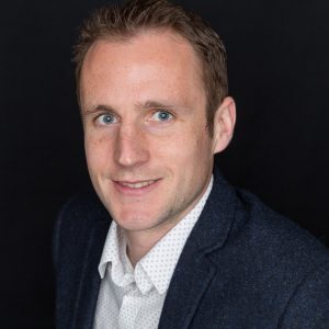 A person with short hair, wearing a dark blazer and white shirt, smiles against a black background.